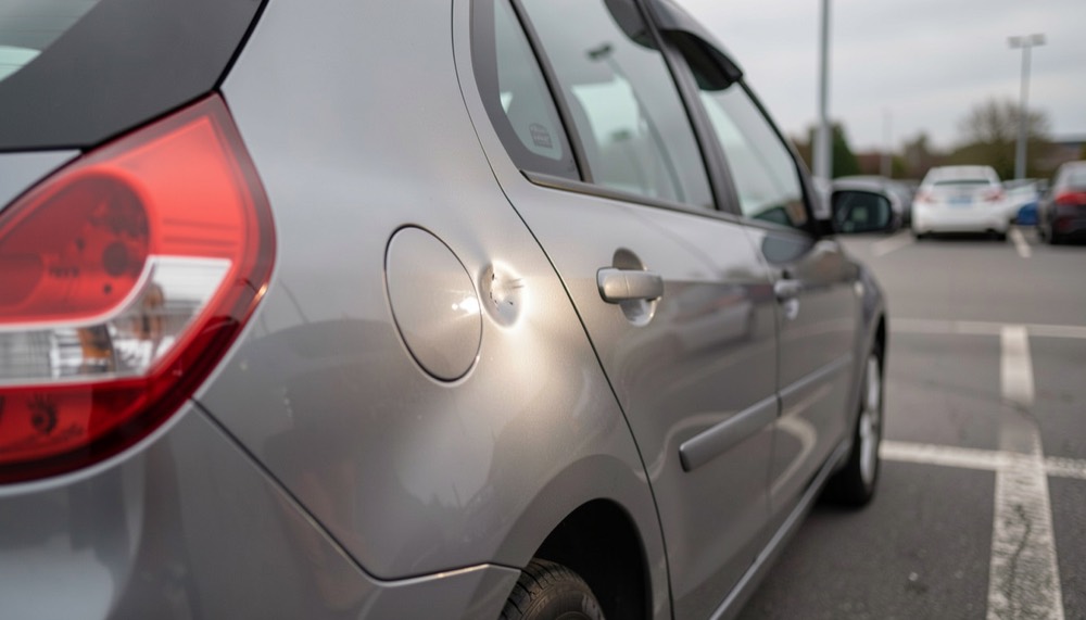 Portière arrière d’une berline grise garée sur un parking, une petite bosse ronde révélée par les reflets d’une lampe, lumière du jour voilée